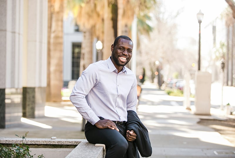 An African American man smiles at the camera while sitting on a sidewalk bench.